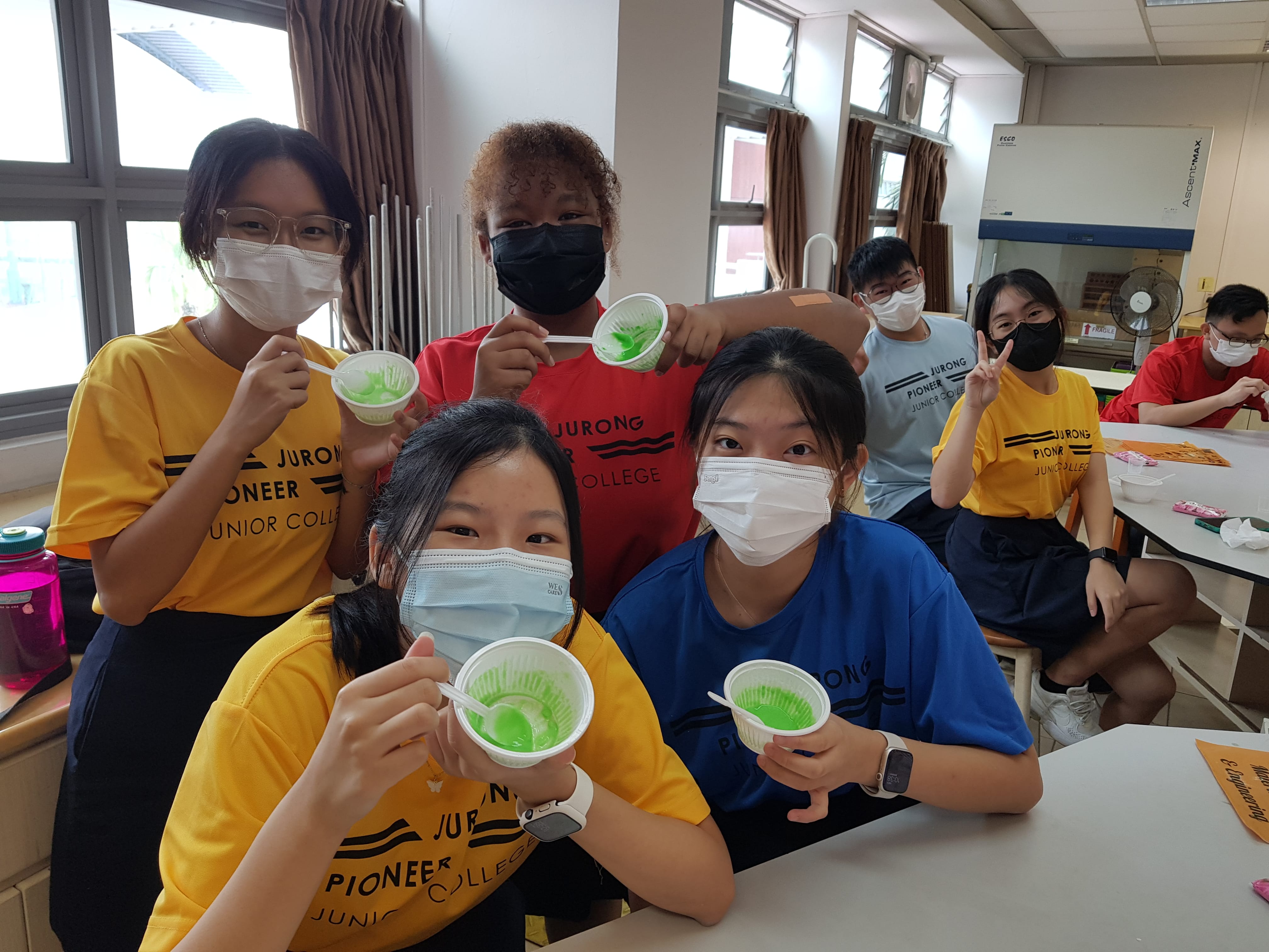 Students in Jurong Pioneer Junior College shirts and face masks hold cups of green jelly in a classroom.